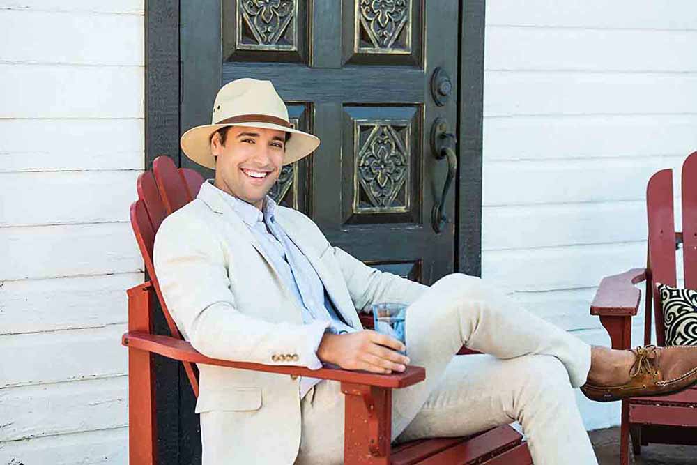 Man is sitting on adirondack chair in tan suit wearing a straw panama hat 
